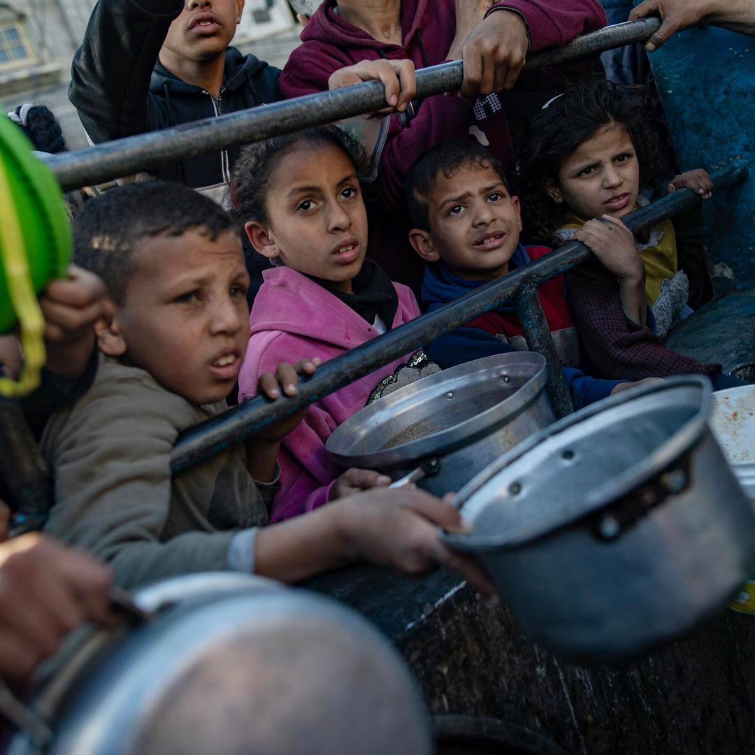 Several Palestinian children with distressed expressions hold out various metal pans, some with handles, some appear to have been from rice cookers, desperately waiting for food. The image makes it clear there are even more out of frame.